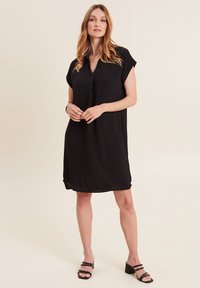 Abito nero a maniche corte con scollo a V, vestibilità ampia e spacco laterale. Tessuto morbido, lunghezza al ginocchio, abbinato a sandali neri con tacco a blocco.