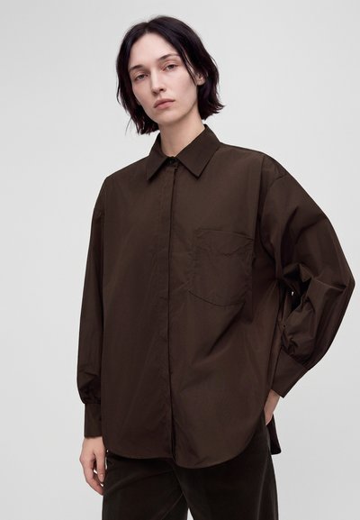 Jeune personne aux cheveux courts et foncés porte une chemise marron foncé ample à boutons avec une poche frontale et des manches longues, debout avec une main dans la poche.