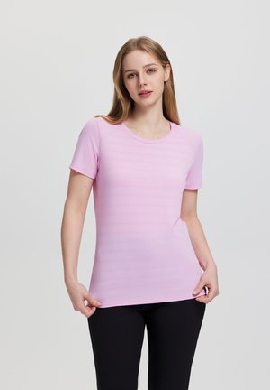 Jonge vrouw met lang blond haar, gekleed in een lichtroze shirt met korte mouwen en zwarte broek, staand en het zoom van het shirt vasthoudend.