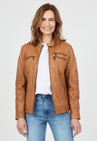Bruine leren motorjack met ritssluitingen in de zakken, geribbelde accenten op de schouders en een getailleerd ontwerp, gedragen over een wit T-shirt en blauwe spijkerbroek.
