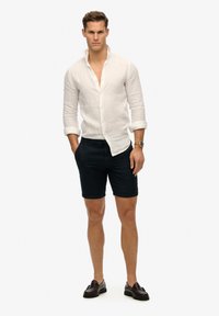 Weißes Leinenhemd mit hochgekrempelten Ärmeln, kombiniert mit dunklen Shorts und braunen Slippern. Das Outfit zeichnet sich durch ein lässiges, aber dennoch tailliertes Design aus.