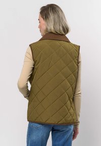 Olivengrønn quiltet vest med brun kordfløyelskrage, med et diamantmønster og ermeløs design. Brukes over en lysfarget topp.