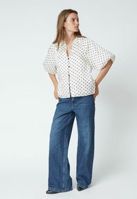 Weiße Kurzarmbluse mit schwarzen Punkten, Knopfverschluss vorne, kombiniert mit hellblauen weit geschnittenen Jeans. Glatte Stoffqualität, lässige Passform.