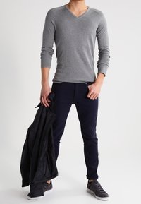 T-shirt gris à manches longues avec col en V, associé à un pantalon slim bleu marine foncé et des baskets gris foncé, tenant une veste noire.