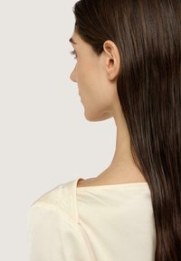 Helles beiges Oberteil mit lockerem Sitz, das eine glatte Textur und einen weiten Ausschnitt aufweist, von hinten gezeigt mit langen, glatten braunen Haaren.