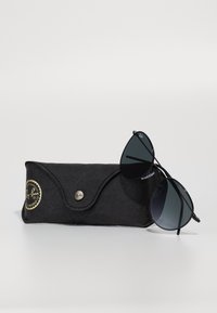 Ochelari de soare Ray-Ban cu lentile negre, în formă de picătură și rame subțiri din metal, așezați pe un etui moale negru cu un logo auriu și închidere tip clapă.
