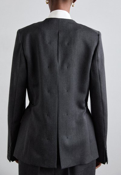 Szürke, testre szabott blazer, struktúrált sziluettel, hátul egyetlen szellőzőnyílással és fehér gallérral. Az anyag finom textúrával és fénylő felülettel rendelkezik.