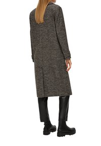 Lange zwart-wit houndstooth jas met een kraag, met een rechte snit en middenlange mouwen, gecombineerd met zwarte leren broeken en laarzen.