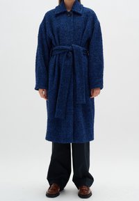 Blå strukturerad kappa med bälte, oversized passform, stor krage och knappstängning. Kombinerad med svarta byxor och bruna skor.