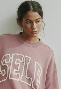 Hele roosa pusa ümar kaelusega, millel esiküljel on valged reljeefsed tähed, mis moodustavad sõna "SELF". Sile tekstuur.