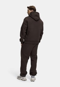 Brun hettegenser og matchende joggebukser, laget av teksturert stoff, med en stor hette, ribbede mansjetter og avslappet passform. Joggesko fullfører antrekket.