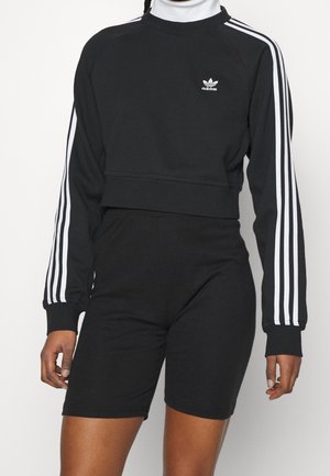 Czarny krótki top Adidas z długim rękawem i krótkie spodenki rowerowe z wysokim stanem, z białymi paskami na rękawach, noszone przez osobę o splecionych włosach.