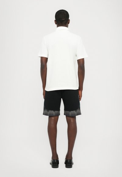 Camisa polo branca combinada com calções pretos com um remate em padrão zig-zag. Os calções são lisos e a polo tem um design de gola clássica.