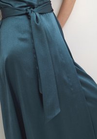 Teal sametimekkoinen puku, jossa on solmittava vyö vyötäröllä. Sen pinnalla on sileä rakenne ja hienovarainen kiilto. Kangas laskeutuu elegantisti.