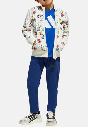 Giacca bianca con zip e motivi grafici colorati, t-shirt blu con logo, pantaloni sportivi blu e scarpe da ginnastica nere con accenti bianchi.
