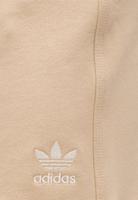 Μπεζ ύφασμα με λευκό λογότυπο τριφύλλι Adidas και κείμενο κεντημένα κοντά σε μια κάθετη ραφή.