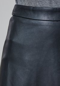 Jupe en cuir noir avec une texture lisse et une taille pliée. La surface présente un léger motif de grain.