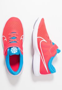 Buty sportowe z jasnoróżową siateczkową cholewką, niebieskim akcentem, białym logo w formie swooshu i rzepem. Szara gumowa podeszwa o teksturowanym chwycie.