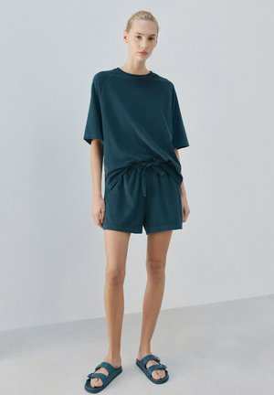 Donna in piedi all'interno, indossa una maglietta a maniche corte color verde acqua scuro, pantaloncini abbinati con coulisse e sandali slid blu, su uno sfondo grigio uniforme.