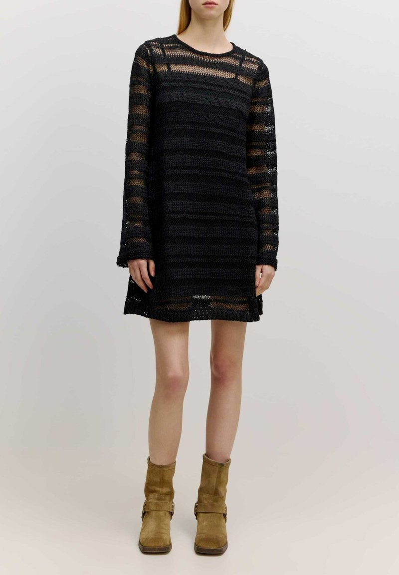 Schwarz gestricktes Kleid mit langen Ärmeln, gestreiftem Design und rundem Ausschnitt. Getragen mit hellbraunen Stiefeletten aus Wildleder.