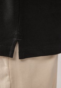 Primer plano del dobladillo de una camisa negra con abertura lateral sobre pantalones beige, destacando la textura de la tela y los detalles de costura de la prenda.
