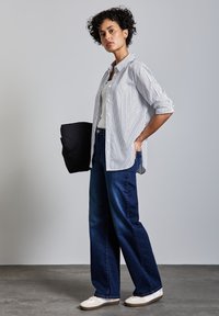 Camicia a righe in bianco e blu navy, abbinata a jeans a gamba larga blu scuro e sneakers bianche. Tiene una borsa nera. Texture del pavimento visibile.
