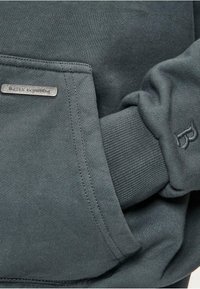 Mørkegrøn sweatshirt med ribbede manchetter og en syet lomme med et metallogo-tag. Stoffet har en glat og blød overflade.