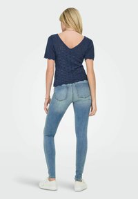 Jūras zilas adītas krekls ar īsām piedurknēm un V formas aizmuguri, apvienots ar gaiši zilām šaurām biksēm un baltām sporta apaviem. Tekstūrēts, ikdienišķs dizains.