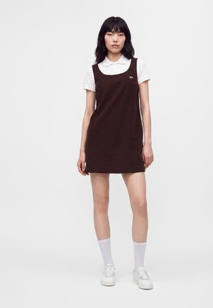Donna che indossa un vestito senza maniche marrone scuro sopra una camicia bianca con collo, calzini bianchi e scarpe da ginnastica bianche, in piedi davanti a uno sfondo bianco uniforme.