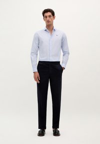 Lichtblauw overhemd met lange mouwen en een klein logo op de borst, gecombineerd met donkere marineblauwe broek en zwarte schoenen.