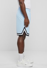 Lyseblå sports-shorts med sort og hvid chevron-detaljer, elastisk talje, og en glat overflade, parret med hvide sneakers.