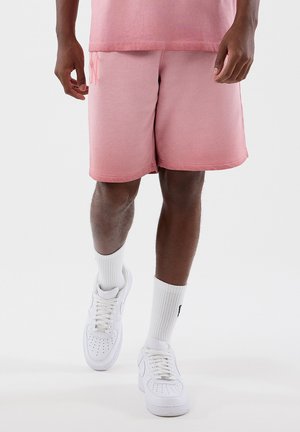 Uomo che cammina con pantaloncini rosa, calzini bianchi alla caviglia e sneaker bianche su uno sfondo chiaro uniforme.