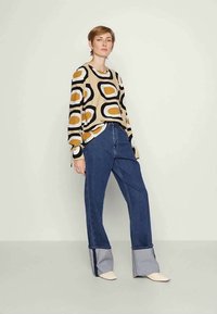 Suéter bege de mangas compridas apresenta padrões ovais pretos e dourados; combinado com jeans de denim, azuis com punhos cinza em contraste e um corte descontraído.