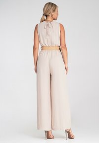 Jasno beżowy kombinezon z zawiązywaniem na plecach, szerokimi nogawkami i plecionym paskiem. Gładka tekstura z luźnym krojem. Widoczne buty na obcasie.