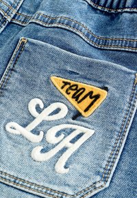 Tascha in denim blu con lettere bianche ricamate "LA" e una piccola etichetta gialla con il testo nero "team".