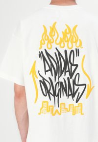 Balta kokvilnas T-kreklā ar lielu melnu un dzeltenu grafisku tekstu un liesmām uz muguras, kurā ir frāze "adidas Originals."