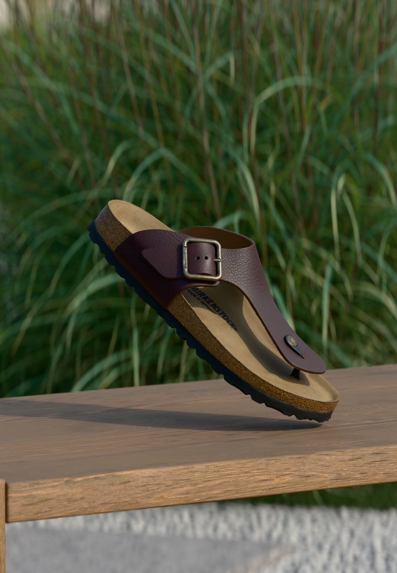 Καφέ δερμάτινο σανδάλι τύπου σφήνα με ρυθμιζόμενο αγκράφα σε ξύλινη επιφάνεια, με θολά πράσινα φυτά στο φόντο.