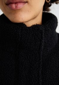 Jachetă din fleece neagră cu un guler înalt și texturat. Prim-plan care arată materialul moale și detaliile cusăturii, cu buzele parțial vizibile.