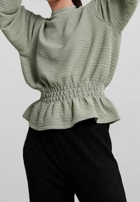 Světle zelený texturovaný top s nařaseným pasem a rozšířeným peplum lemem, kombinovaný s černými žebrovanými kalhotami. Dlouhé rukávy a kulatý výstřih.