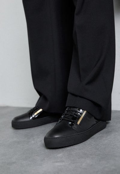 Czarne skórzane sneakersy z błyszczącymi akcentami, teksturowanymi sznurówkami i detalem w postaci zamka; zestawione z ciemnymi, luźnymi spodniami na szarym podłożu.