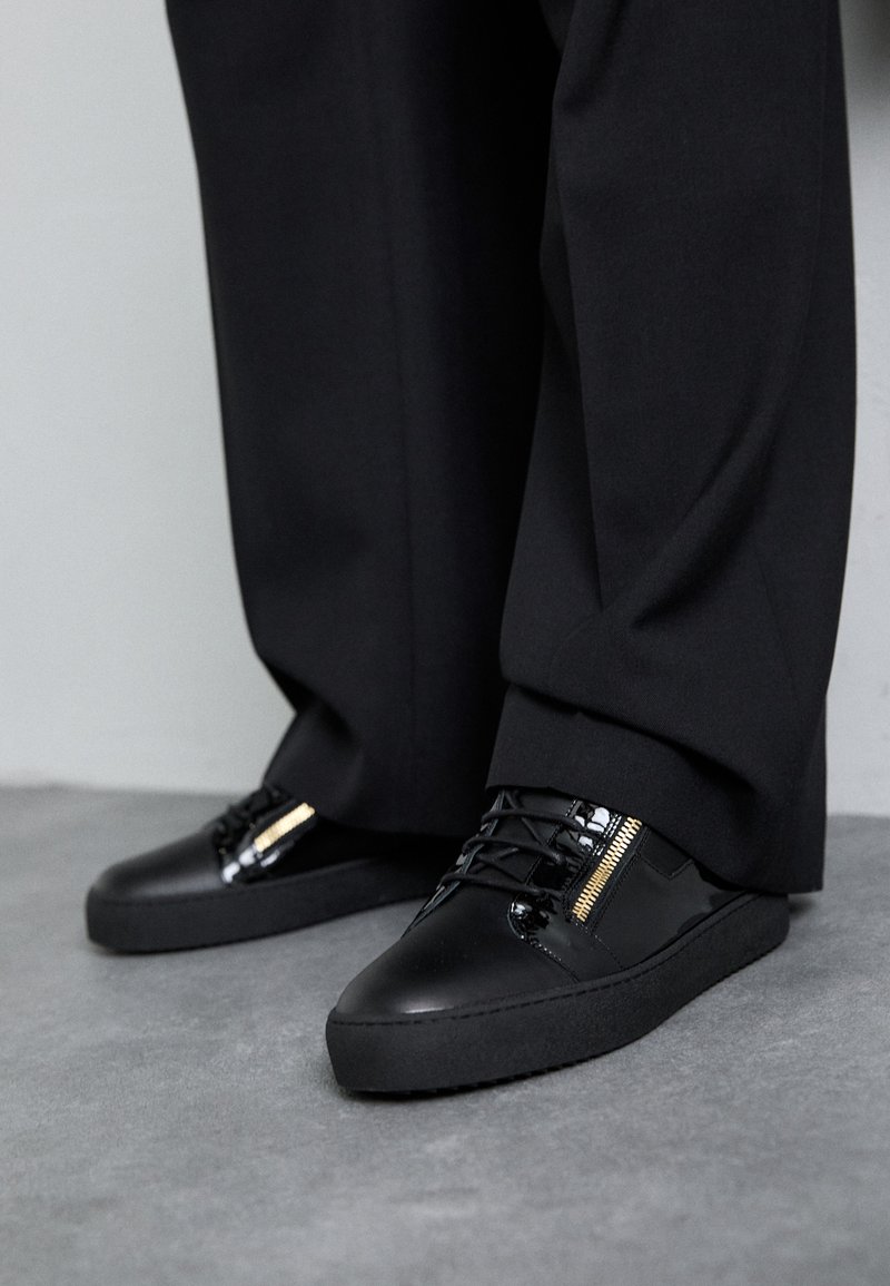 Sapatilhas pretas em couro com acabamentos brilhantes, atacadores texturizados e um detalhe em fecho; combinadas com calças escuras e largas num chão cinza.