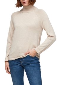 Femme portant un pull à col roulé beige à manches longues et un jean bleu avec des boutons ressemblant à des perles sur la couture latérale.
