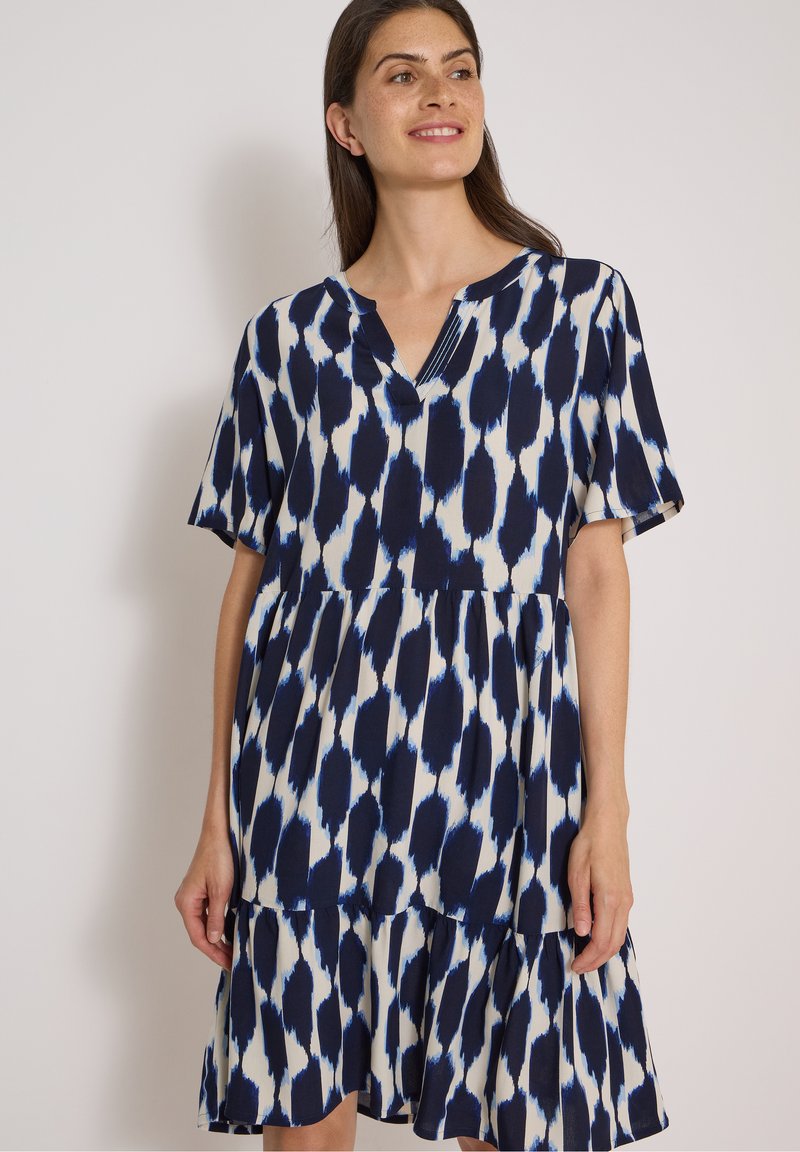 Femme souriante, portant une robe à manches courtes avec un motif abstrait bleu marine et blanc, debout devant un fond clair uni.