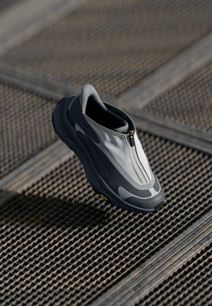 Baskets modernes à enfiler avec fermeture éclair à l’avant, dessus gris et blanc, semelle noire, présentées flottant au-dessus d’une surface métallique texturée.