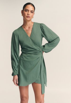 Green wrap dress in textured fabric, featuring a deep V-neck, long sleeves, and an asymmetrical hem with a tied detail at the waist.