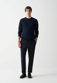Marineblauer Strickpullover mit gerippten Ärmelbündchen und Ausschnitt, kombiniert mit dunklen Hosen mit elastischem Bund und seitlichen Taschen, getragen mit schwarzen Schuhen.