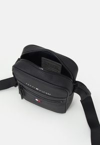 Svart lærcrossbody veske med glidelås, frontlomme og stropp; har preget logo og tricolor aksent nær bunnen.