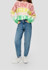 Person som bär en färgglad randig tröja med texten "Roligt är där du är", blå högmidjade jeans, vita sneakers, med händerna i fickorna.