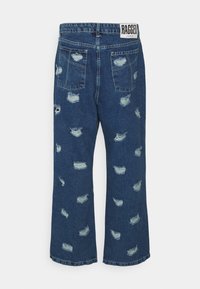 Jeans de ganga azuis em estilo flare com detalhes desgastados, apresentando múltiplos rasgos e remendos desfiados. A etiqueta exibe "RAGGED" na cintura.