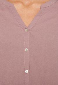 Gros plan sur un chemisier mauve à col en V, avec trois boutons blancs et des coutures visibles le long de l'encolure et de la patte de boutonnage.